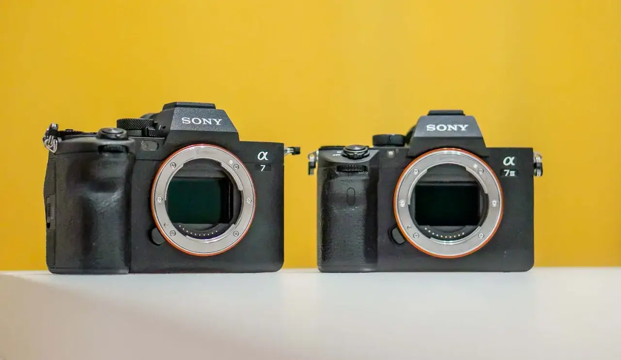 Dwa aparaty Sony Alpha, jeden a7, drugi a7 III, stoją obok siebie na białej powierzchni.