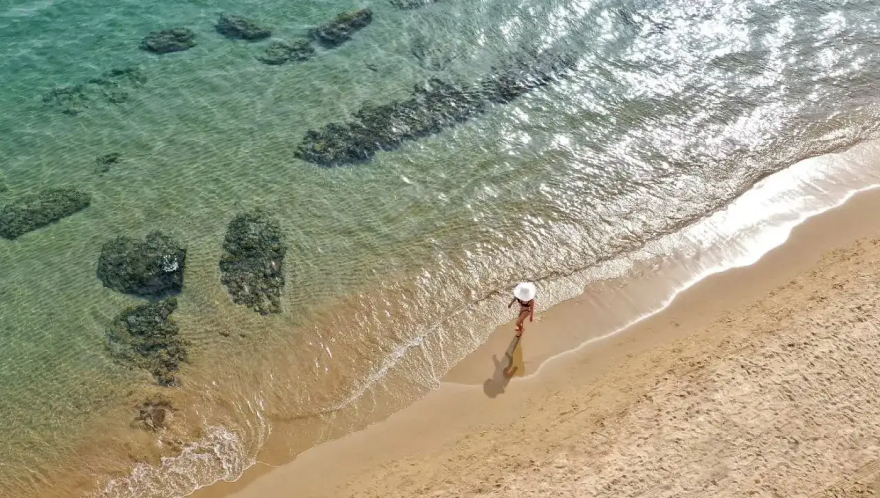 Una donna con cappello bianco cammina sulla spiaggia dorata, con rocce sommerse nell'acqua cristallina. Un'idea per il tuo prossimo viaggio: mare Lazio dove andare.