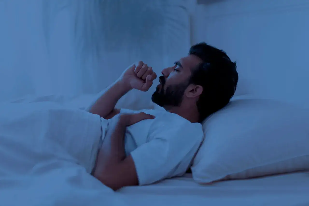 Un homme barbu, allongé dans son lit, se réveille en pleine nuit pour tousser. La lumière bleutée de la chambre accentue son inconfort.