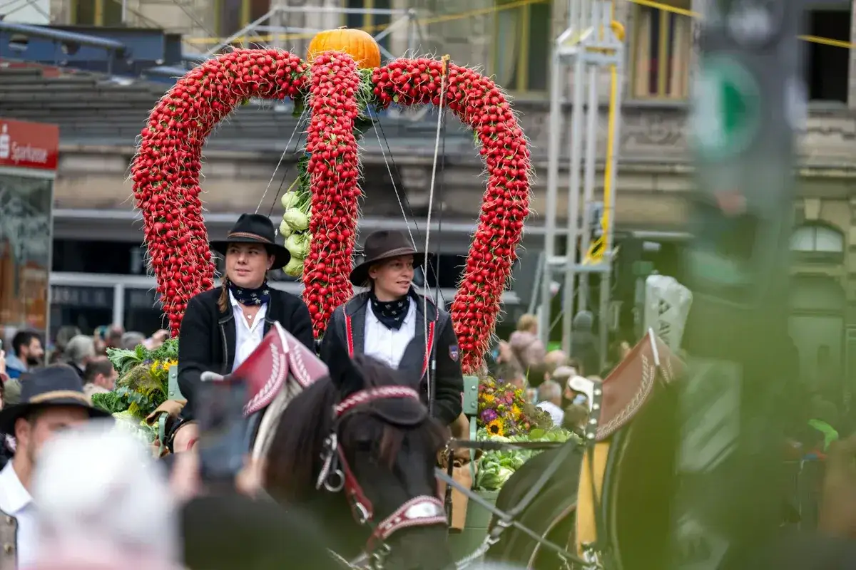 Zwei Frauen in Tracht reiten auf einem Pferd durch eine festliche Parade. Ein riesiges Herz aus Radieschen und ein Kürbis schmücken den Wagen. Das ist typisch für die Kirchweih!