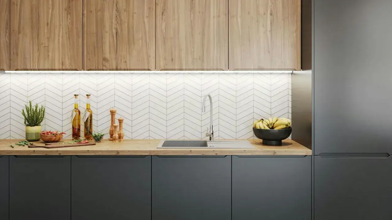 Drewniane wykończenie blatu kuchennego z szarymi szafkami i białym, jodełkowym backsplash. Na blacie miska z bananami, butelki z oliwą i przyprawniki.