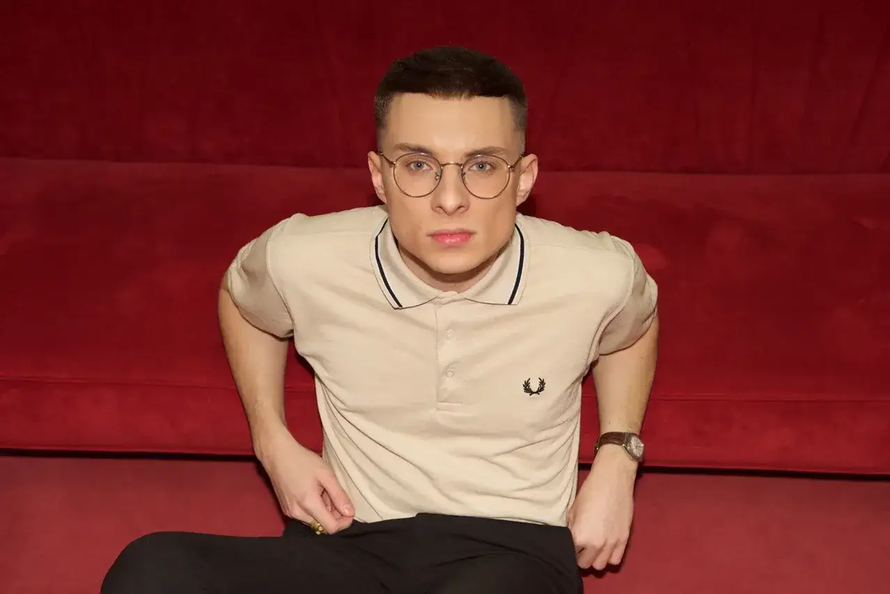 Młody mężczyzna w okularach i beżowej koszulce polo, podobno Ralph Kaminski, siedzi na tle czerwonej kanapy.