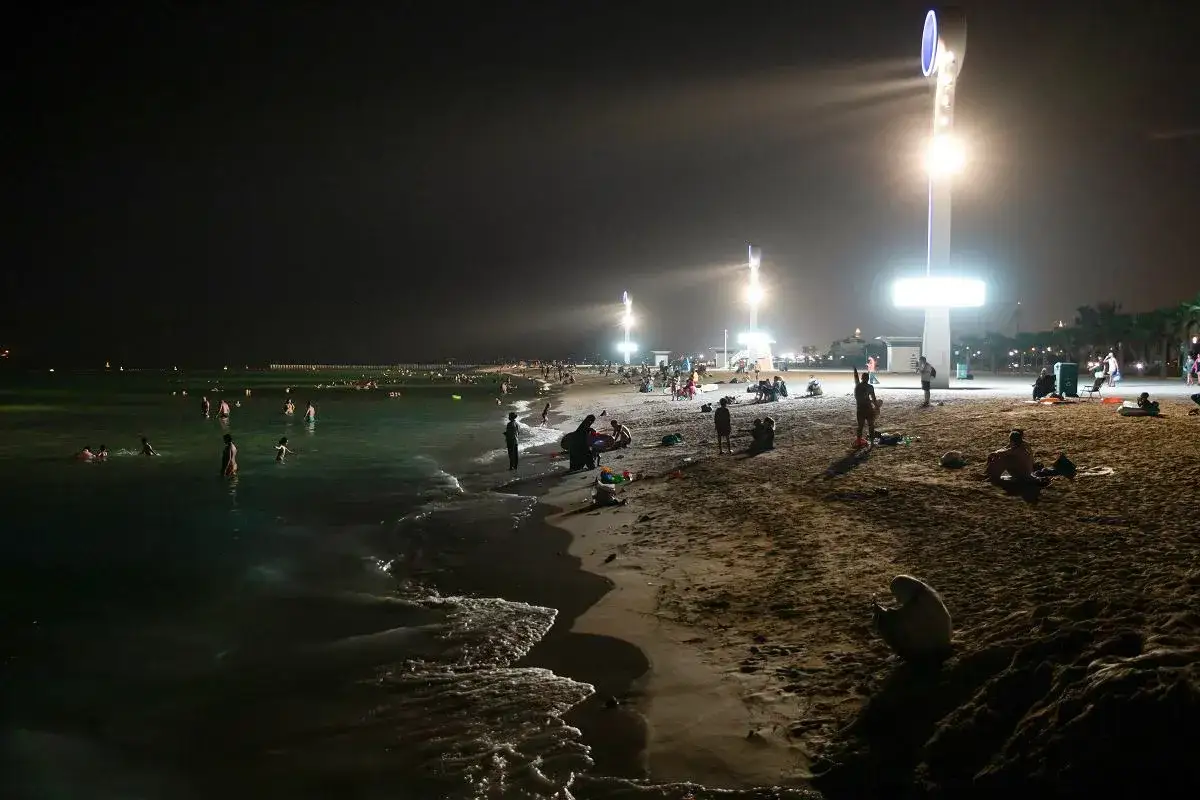 Nocna dubaj plaża tętni życiem. Ludzie kąpią się w morzu i odpoczywają na piasku, oświetleni przez wysokie latarnie.