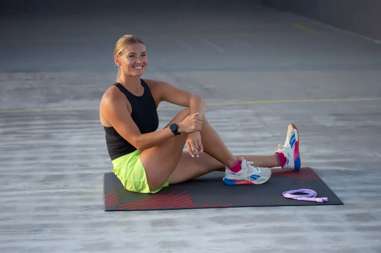 L&auml;chelnde Frau auf Matte, bereit f&uuml;r ein Springseil-Workout. Ein lila Springseil liegt daneben.