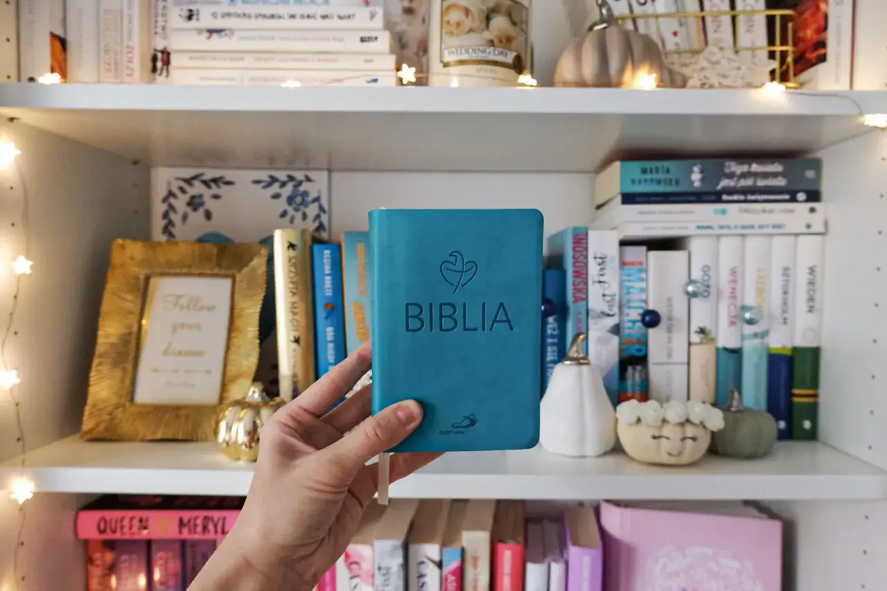 Ręka trzyma turkusową Biblię na tle półki z książkami. Wśród nich widać inne przekłady biblii i ozdoby.