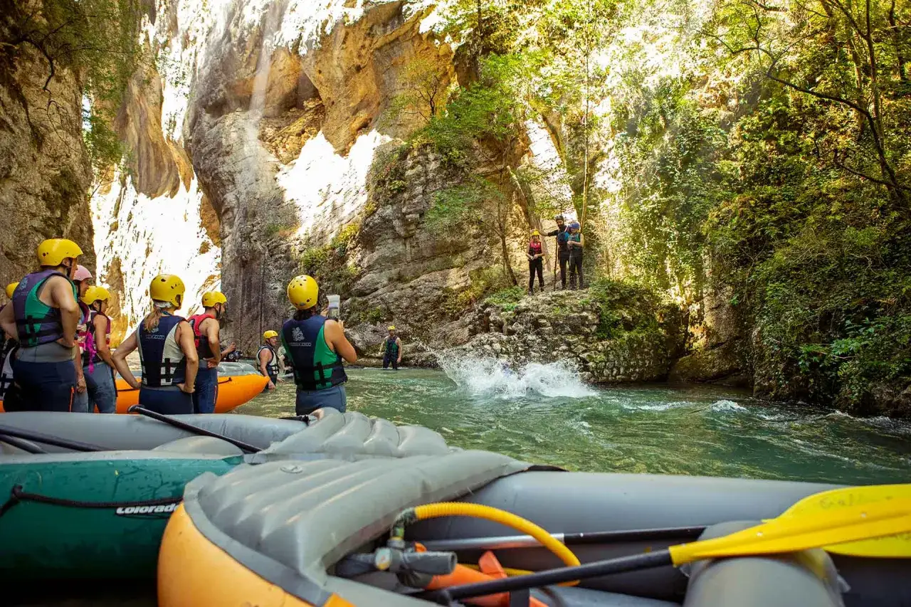 Avventura in rafting tra i suggestivi **paesi della Valnerina**. Un gruppo di persone, equipaggiate con caschi gialli, si prepara a scendere il fiume tra imponenti pareti rocciose e vegetazione lussureggiante.