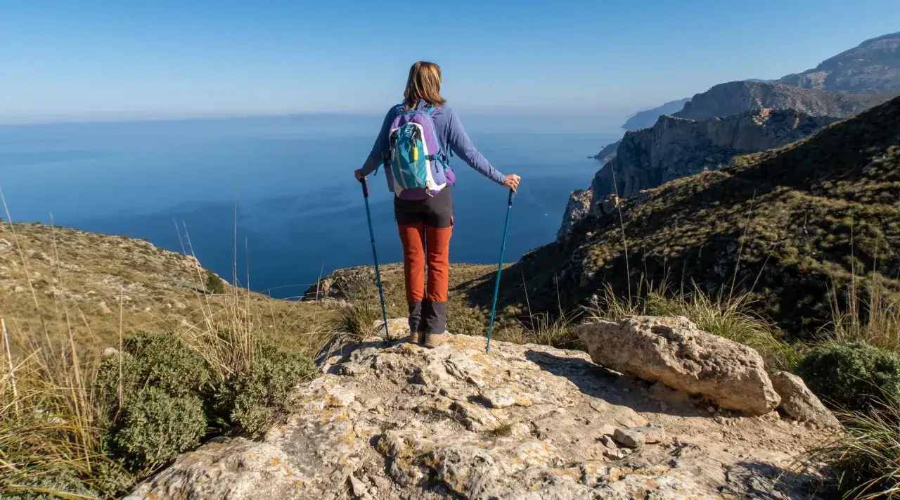 En kvinna med vandringsstavar njuter av utsikten &ouml;ver havet och berg under en vandring p&aring; Mallorca.