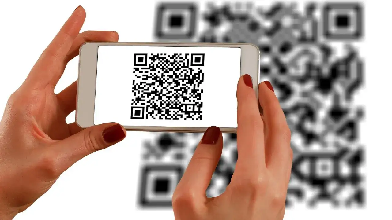 Dwie dłonie trzymają smartfon, na którego ekranie wyświetlony jest kod QR. W tle widać rozmazane kody QR. Dowiedz się, jak zeskanować kod QR ze zdjęcia.