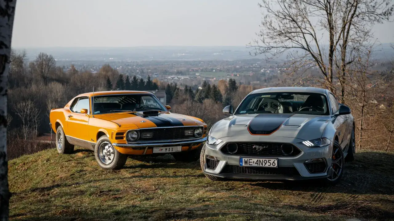 Dwa Fordy Mustang: klasyczny pomarańczowy z lat 70. i nowoczesny szary, oba w wersji **Mustang Mach 1**, stoją na wzgórzu z widokiem na dolinę.