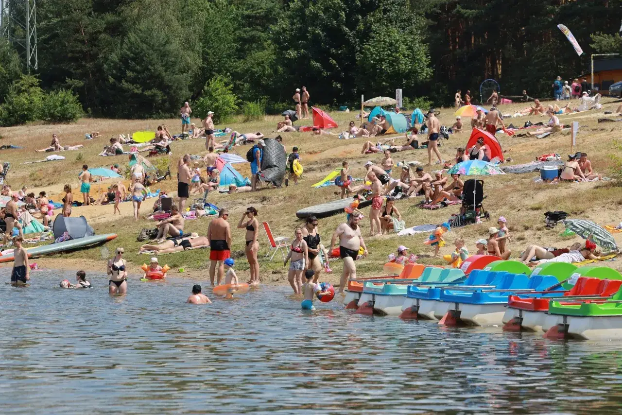Tłum ludzi odpoczywa na piaszczystym **chechle** nad jeziorem, opalając się i kąpiąc.
