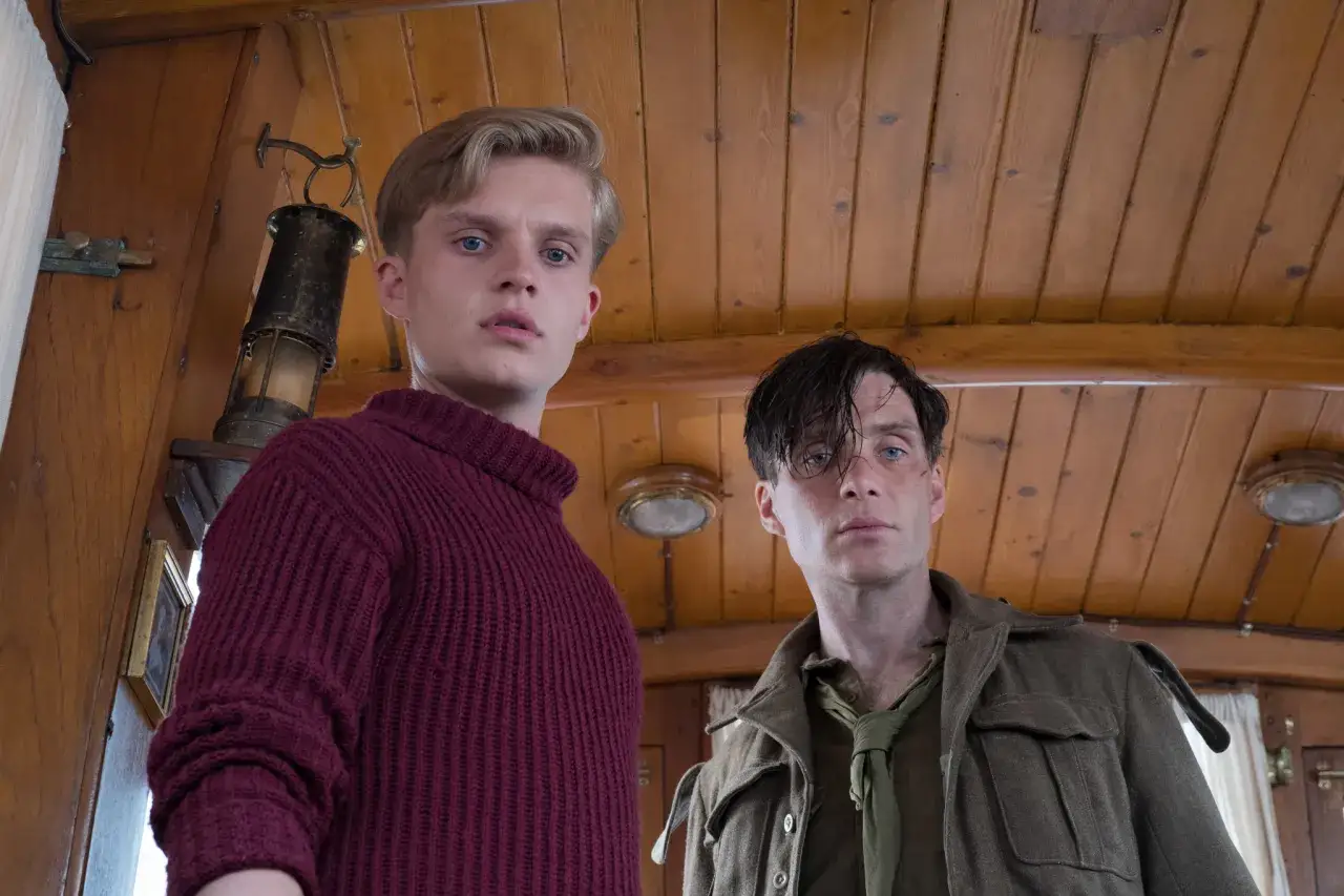 Två män, en ung med blont hår i en vinröd tröja och en äldre med mörkt hår i militäruniform, tittar mot kameran. De befinner sig ombord på ett fartyg, kanske under evakueringen i Dunkirk.
