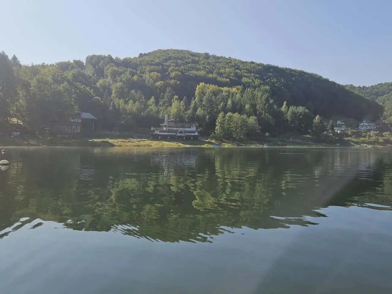 Spokojny poranek nad jeziorem, zalesione wzg&oacute;rza odbijają się w wodzie. Widoczna łososina dolna z zabudowaniami.