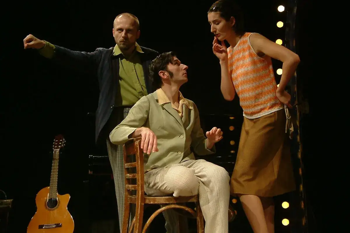 Trzech aktorów na scenie, gitara obok. Mężczyzna w marynarce gestykuluje, siedzący wpatrzony w kobietę w pomarańczowej bluzce. Teatr ciut frapujący.