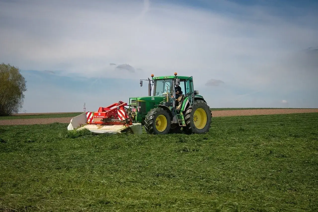 Zielony traktor z zielonymi kołami i czerwonym osprzętem kosi trawę na polu. Traktorzysta pracuje w słoneczny dzień.