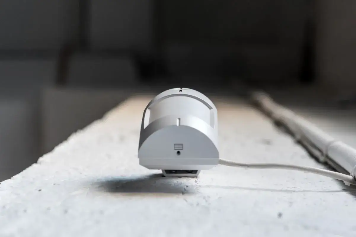 Detector de presencia y movimiento, un dispositivo blanco con cable conectado, montado en una superficie rugosa.