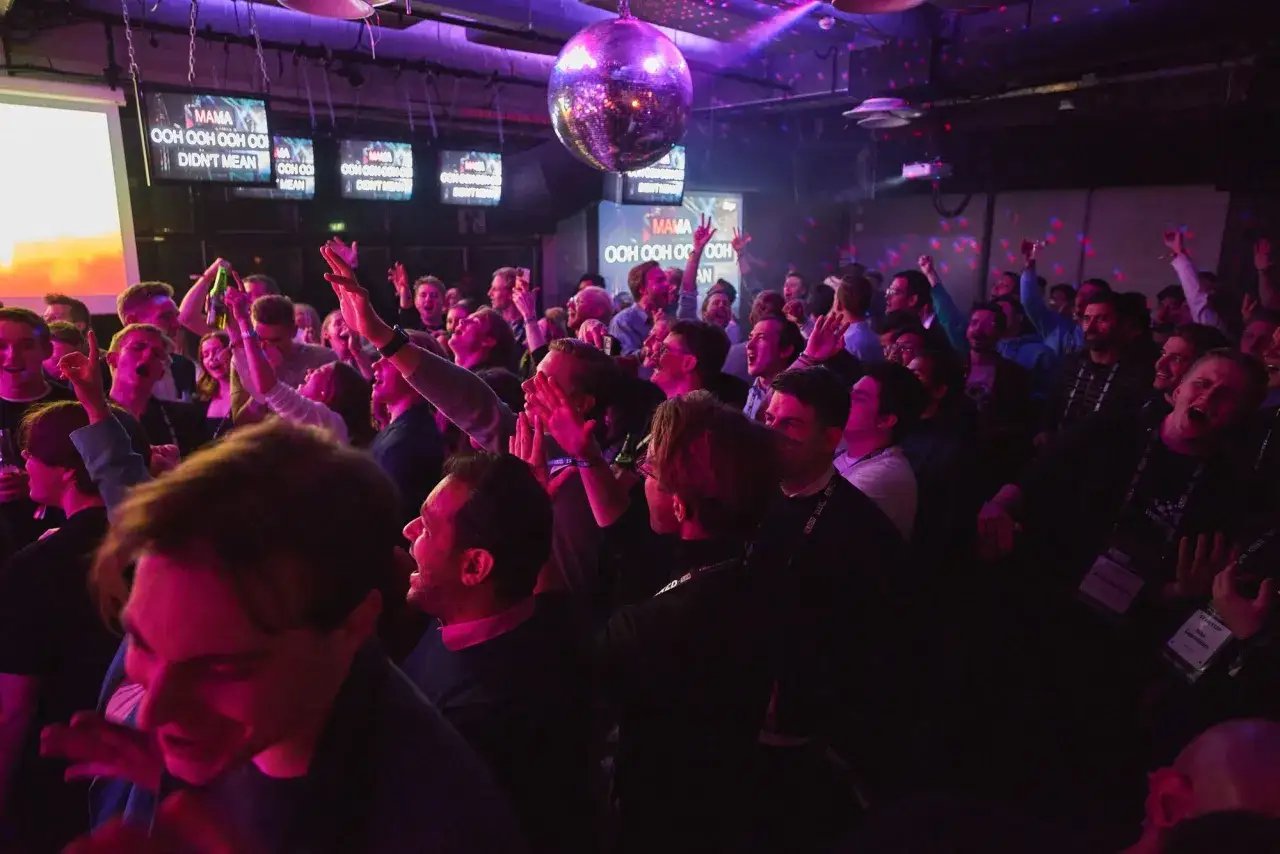 Menschen feiern ausgelassen in einer Karaoke-Bar, singen und tanzen zur Musik. Eine Discokugel funkelt über der Menge.