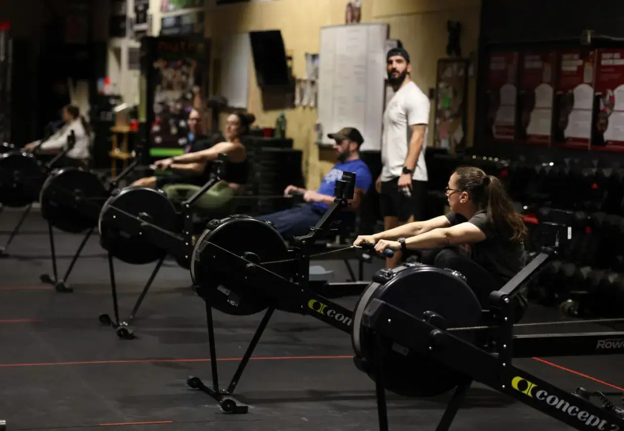 Ludzie ćwiczą na wioślarzach w sali treningowej. To pokazuje, crossfit co to za trening – intensywny i wszechstronny.