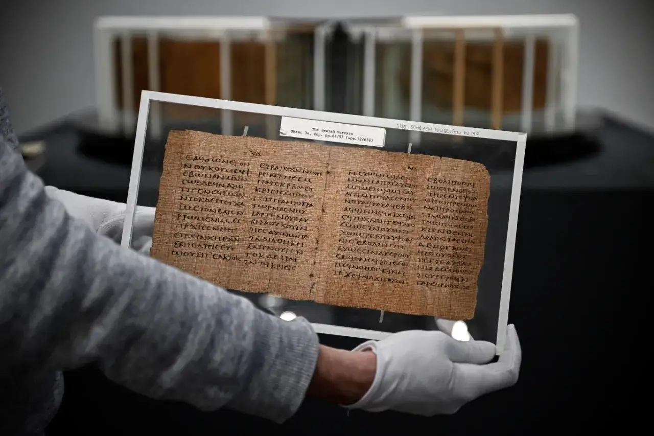 Fragment papirusu z tekstem biblijnym, być może psalm 135, w rękach osoby w białych rękawiczkach.