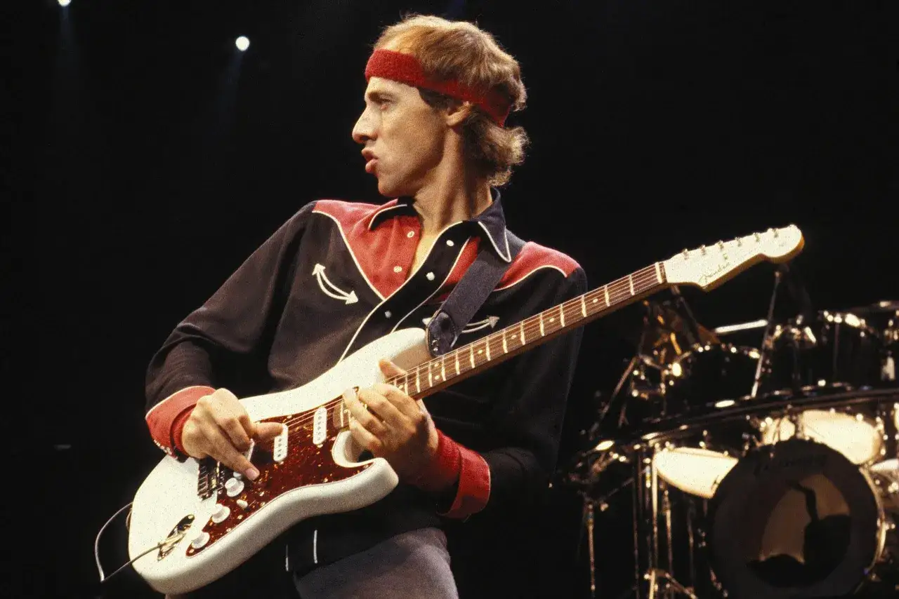 Mark Knopfler w czerwonej opasce na głowie gra na gitarze Fender Stratocaster. W tle widoczna perkusja.
