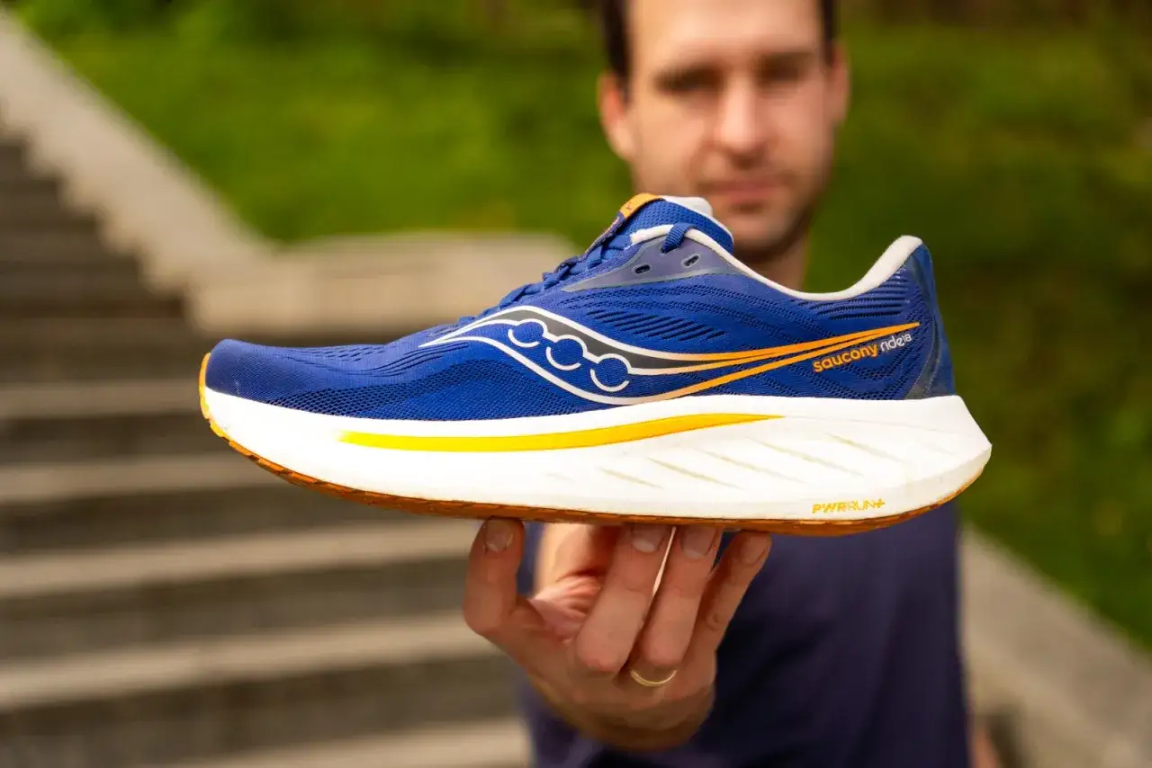 Mężczyzna prezentuje niebieskie buty do biegania Saucony Ride 18 z pomarańczowymi akcentami.