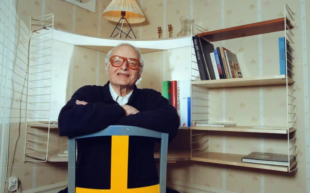 Äldre man med glasögon sitter framför en hylla med böcker, en svensk designklassiker.