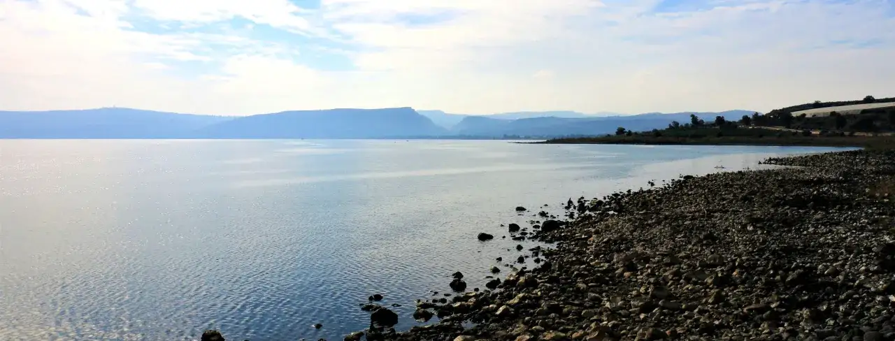 Spokojne wody Jeziora Galilejskiego, gdzie Jezus nauczał w Kafarnaum. Kamienista plaża i góry w oddali.