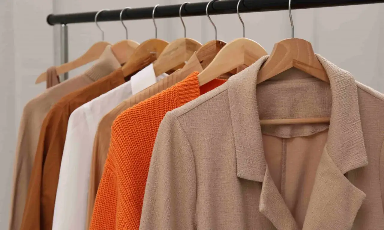 Eine Reihe von Kleidungsst&uuml;cken h&auml;ngt an Kleiderb&uuml;geln. Ein beiger Blazer ist im Vordergrund, gefolgt von einem orangefarbenen Strickpullover, einem wei&szlig;en Hemd, einem braunen Pullover und einem hellbraunen Pullover. Zeit, den Anzug waschen zu lassen.