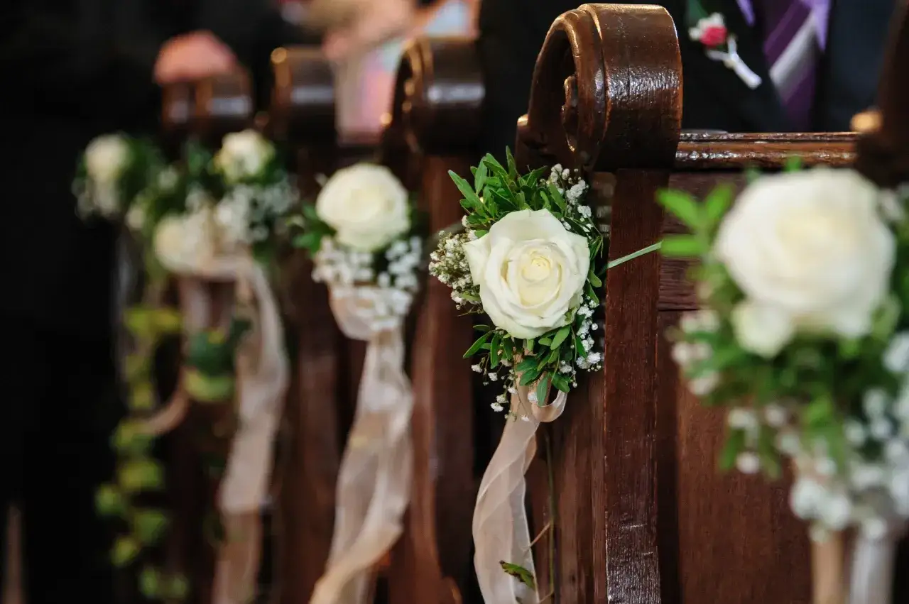 Elegancka dekoracja kościoła na ślub samemu: białe róże z gipsówką i zielenią, ozdobione wstążkami, przy drewnianych ławkach.