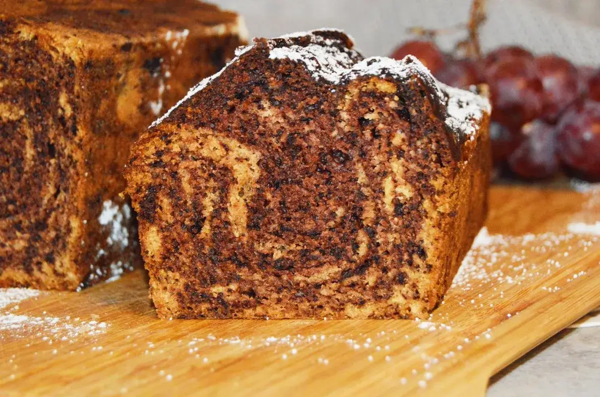Puszysty chlebek bananowy z kakao, opr&oacute;szony cukrem pudrem, kusi apetycznym marmurkowym wzorem. Obok kiść winogron.