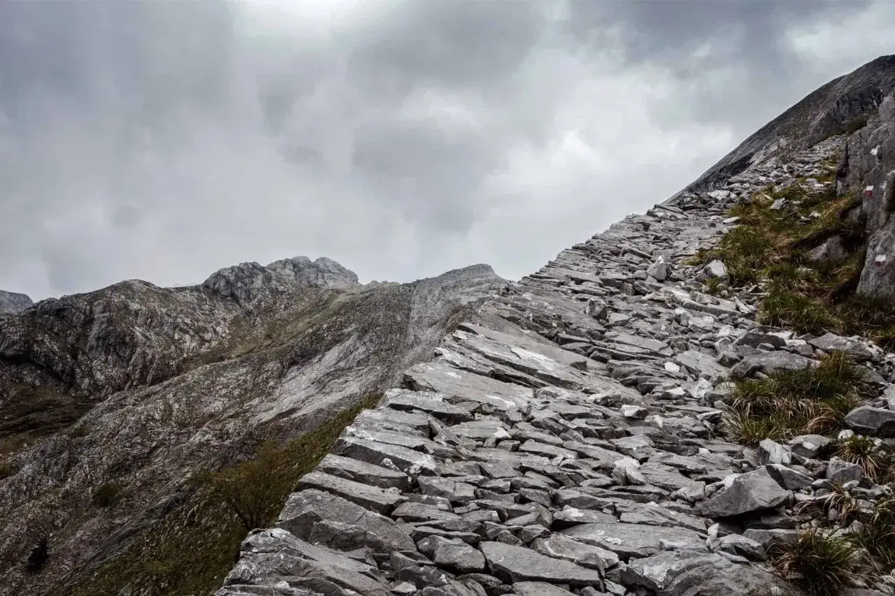 Sentiero roccioso e scosceso su **la via Vandelli**, con montagne grigie e cielo nuvoloso.