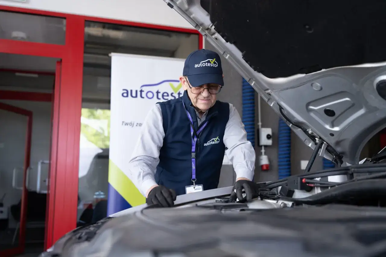 Mechanik w czapce z logo autotesto sprawdza silnik samochodu.