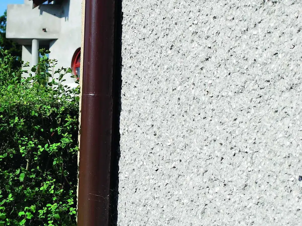 Szara, chropowata ściana z kamyczkami, idealna jako grunt pod farbę elewacyjną. Obok rynna i zielone krzewy.
