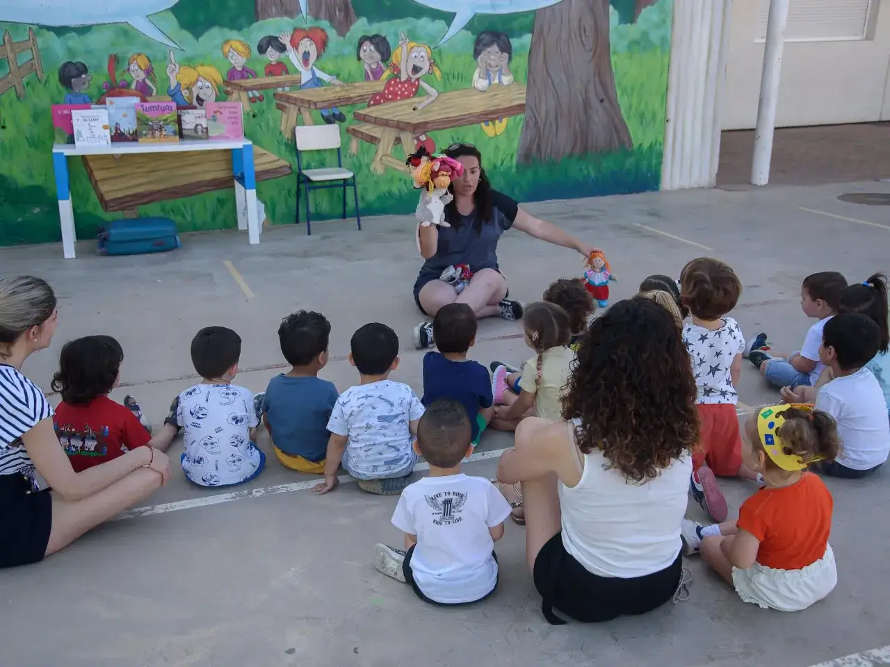Una maestra cuenta cuentos con t&iacute;teres a un grupo de ni&ntilde;os de guarder&iacute;a de 2 a&ntilde;os, sentados en el suelo frente a un mural colorido.