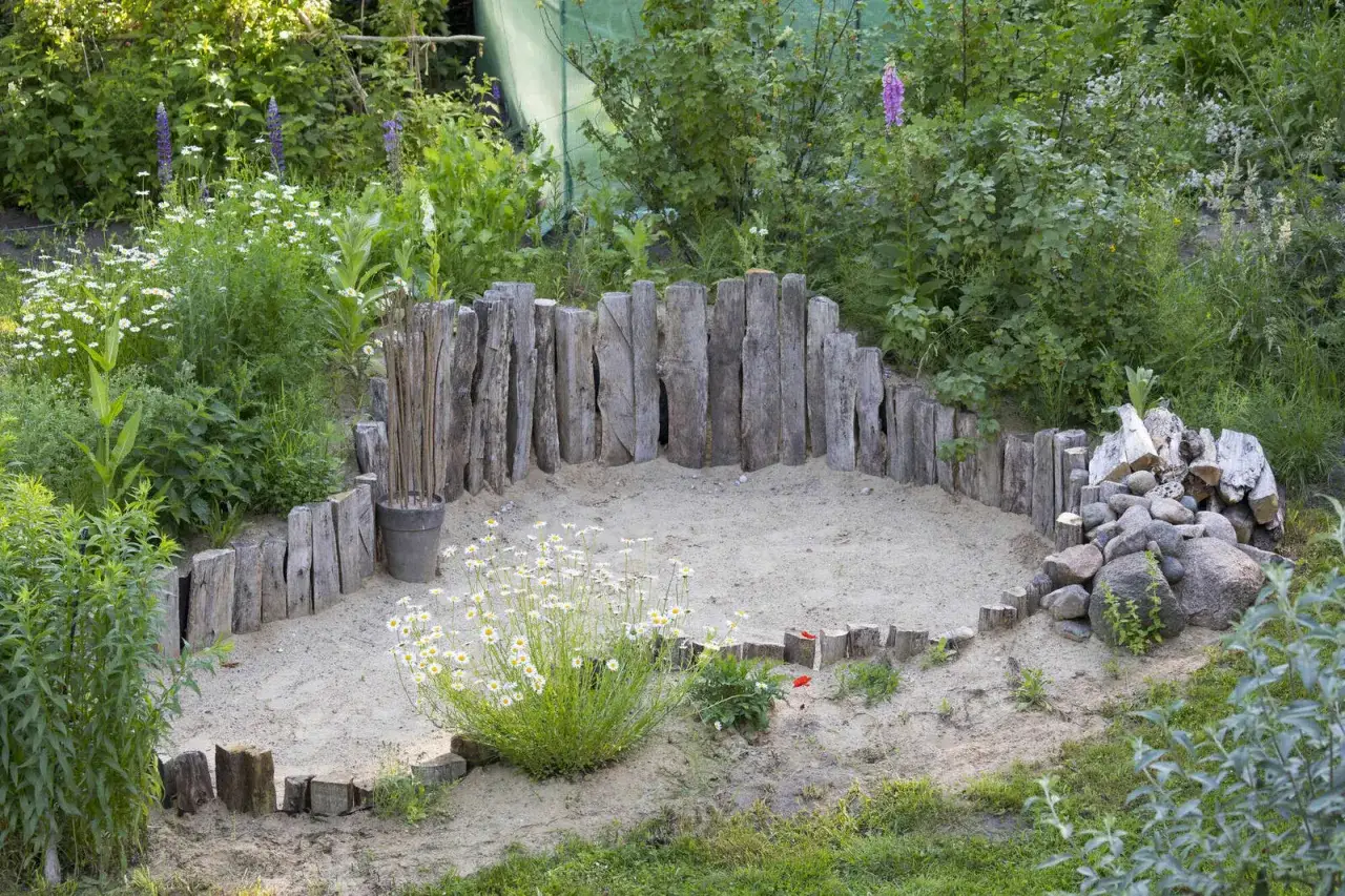 Ein Sandbereich mit Holzstämmen, der als wildbienen nest dient. Blumen und Steine umgeben den Bereich.