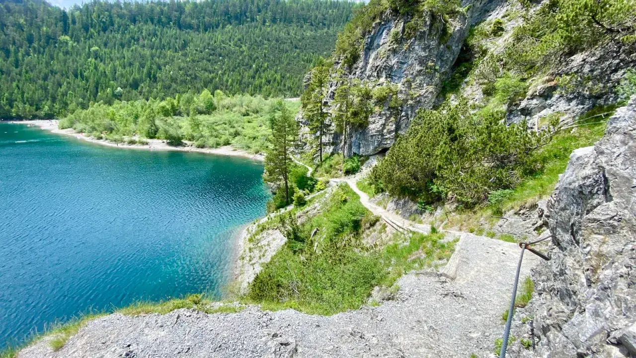 Ein schmaler blindsee trail schlängelt sich an einem steilen Felsen entlang, mit türkisfarbenem Wasser und grünen Wäldern im Hintergrund.