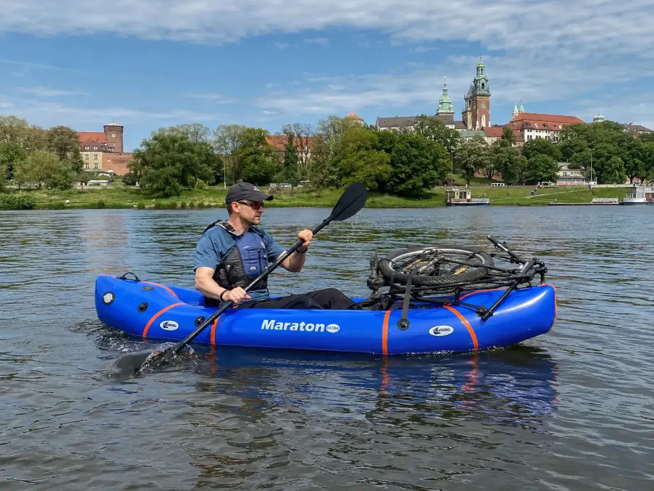 Mężczyzna na dmuchanej łodzi z rowerem z tyłu, uprawiający bikerafting po rzece z widokiem na zamek.