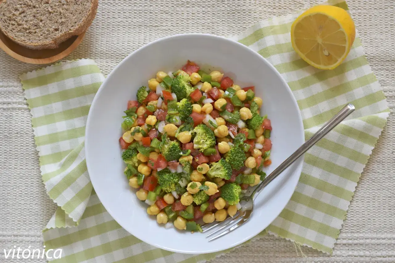 Ensalada garbanzos vibrante con brócoli, tomate y pimiento verde. Servida en un bol blanco con pan y limón al lado.