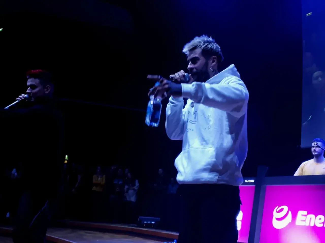 Quebonafide koncert: raper w białej bluzie z kapturem śpiewa do mikrofonu, trzymając butelkę wody. Na scenie widać też innych artystów i publiczność.