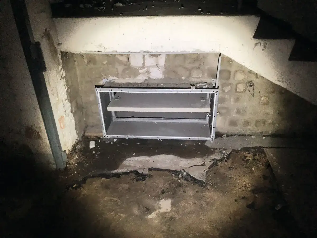 Einbau von Brandschutzklappen in einem alten Gebäude, möglicherweise mit Asbest. Die Klappen sind in einer Wandöffnung montiert, der Boden ist feucht und beschädigt.