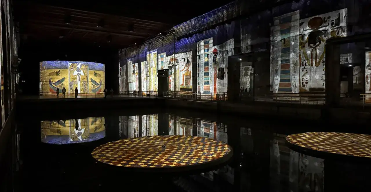 Ancient Egyptian hieroglyphs and figures illuminate the walls and a cylindrical structure at the Bassins des Lumières, reflected in the dark water.