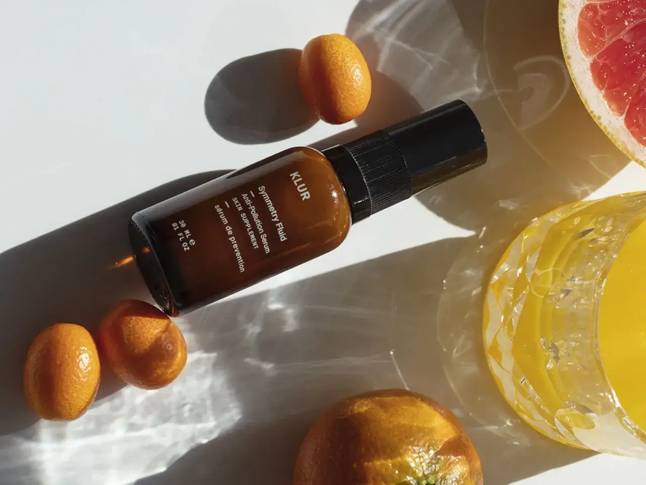 Serum KLUR Symmetry Fluid chroni skórę przed stresem oksydacyjnym. Wokół buteleczki kumkwaty, plaster grejpfruta i szklanka soku.