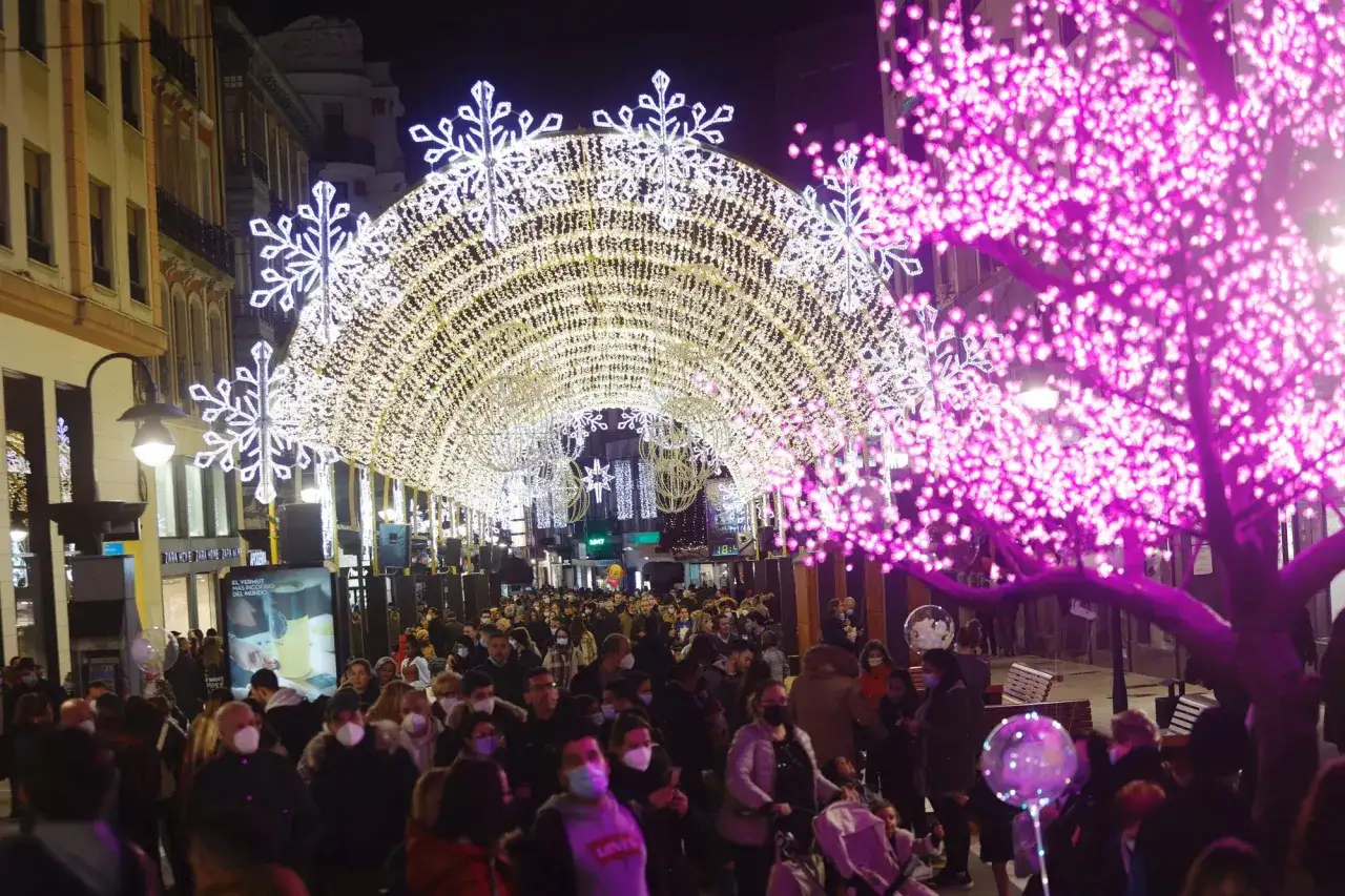 Gente disfrutando del encendido de luces en Oviedo, con un arco de luces blancas y copos de nieve, y un árbol de luces rosas.