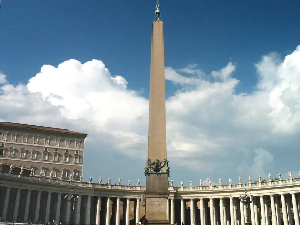 Wielki obelisk na placu Świętego Piotra w Watykanie, otoczony kolumnadą Berniniego, pod błękitnym niebem z białymi chmurami.