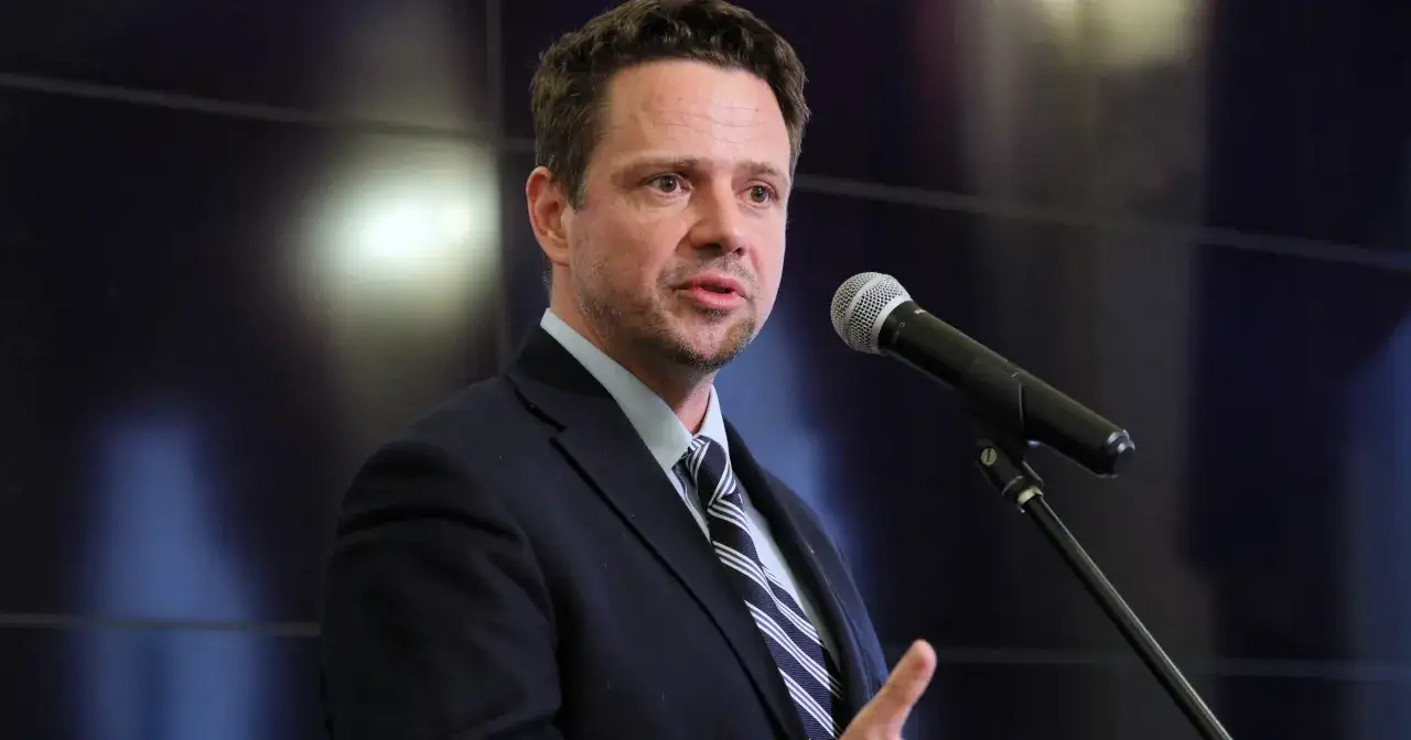 Rafał Trzaskowski, którego wiek budzi zainteresowanie, przemawia do mikrofonu w garniturze.