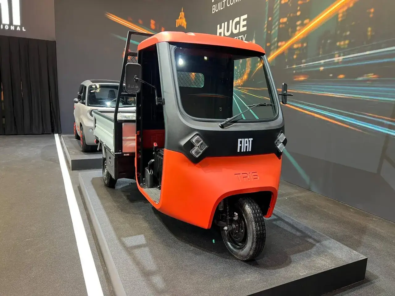 Un veicolo a tre ruote Fiat Tris arancione e grigio, con un cassone aperto, esposto su una pedana.