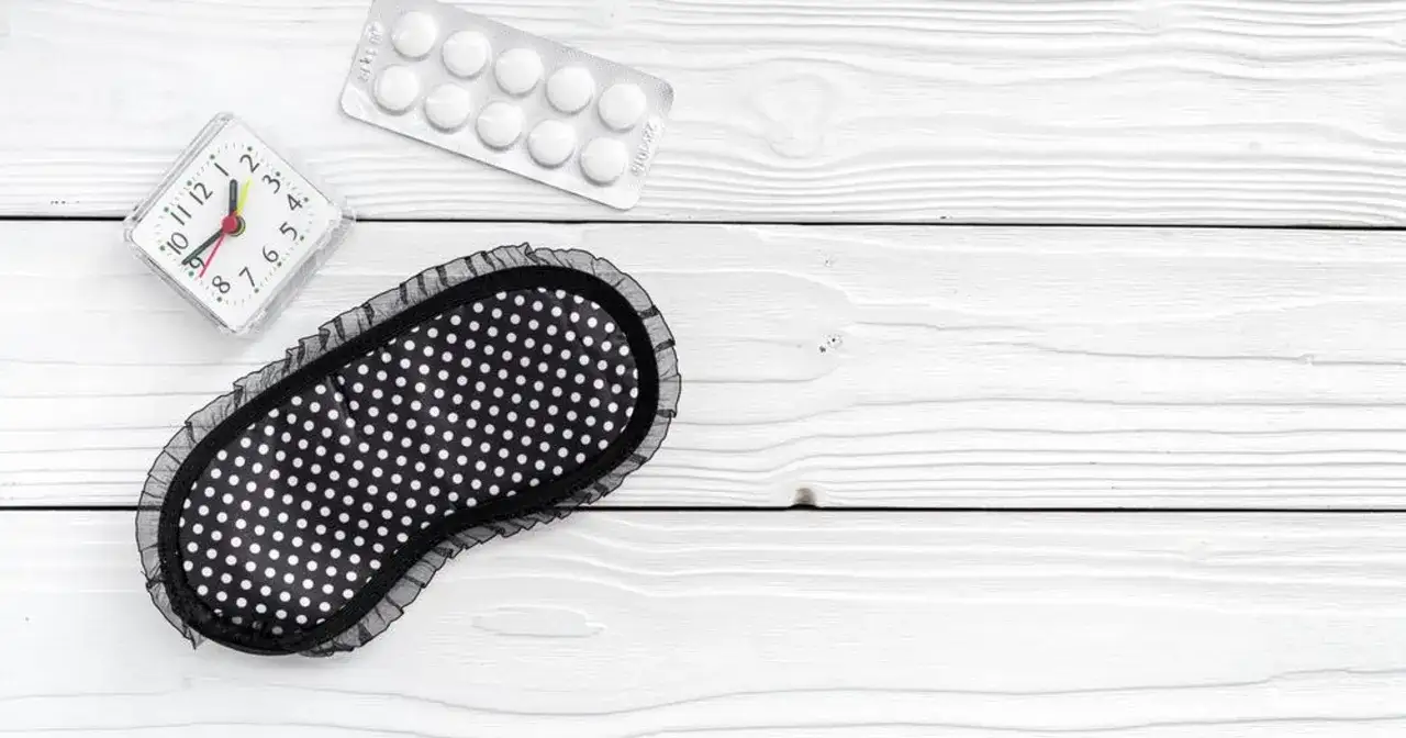 Budzik, maska na oczy i opakowanie tabletek, być może zolpidem, leżą na białym, drewnianym tle.