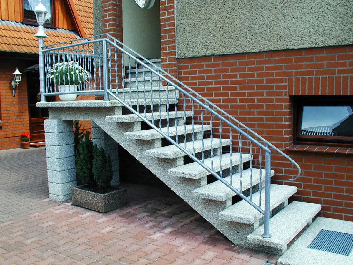 Solidna, zewnętrzna klatka schodowa w domu jednorodzinnym, z kamiennymi stopniami i metalową balustradą, prowadzi do wejścia.