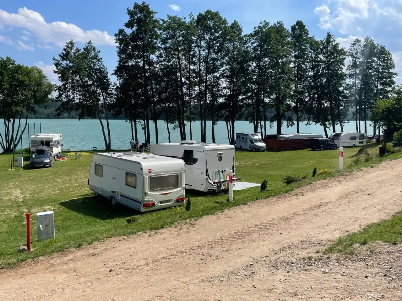 Idealne miejsce, gdzie pod namiot nad jezioro. Kemping z przyczepami i kamperami na zielonej trawie, tuż przy turkusowej wodzie i drzewach.
