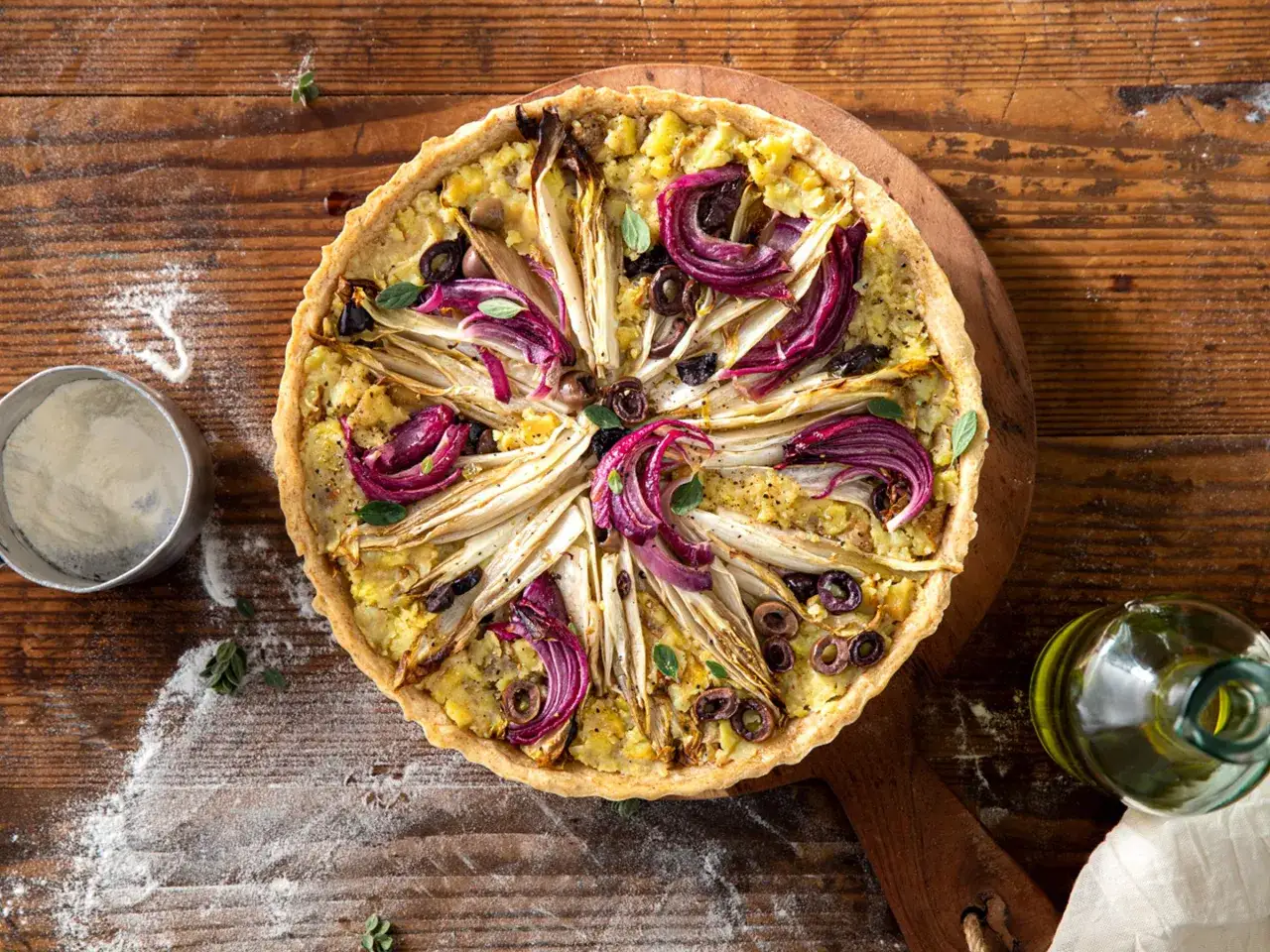 Torta salata con cicoria, cipolla rossa e olive su pasta brise, accanto a una bottiglia di olio.