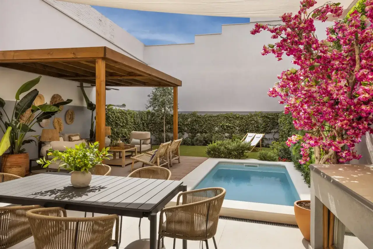Un patio moderno con piscina, zona de comedor exterior y un rincón chill-out bajo una pérgola de madera. Flores rosas vibrantes añaden color.